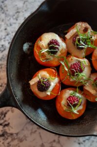 apricots face up in cast iron skillet with brie and blackberries in the hollow of each, garnished with slivered basil