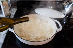 wine is being poured into a dutch oven containing rice, shallot, and garlic